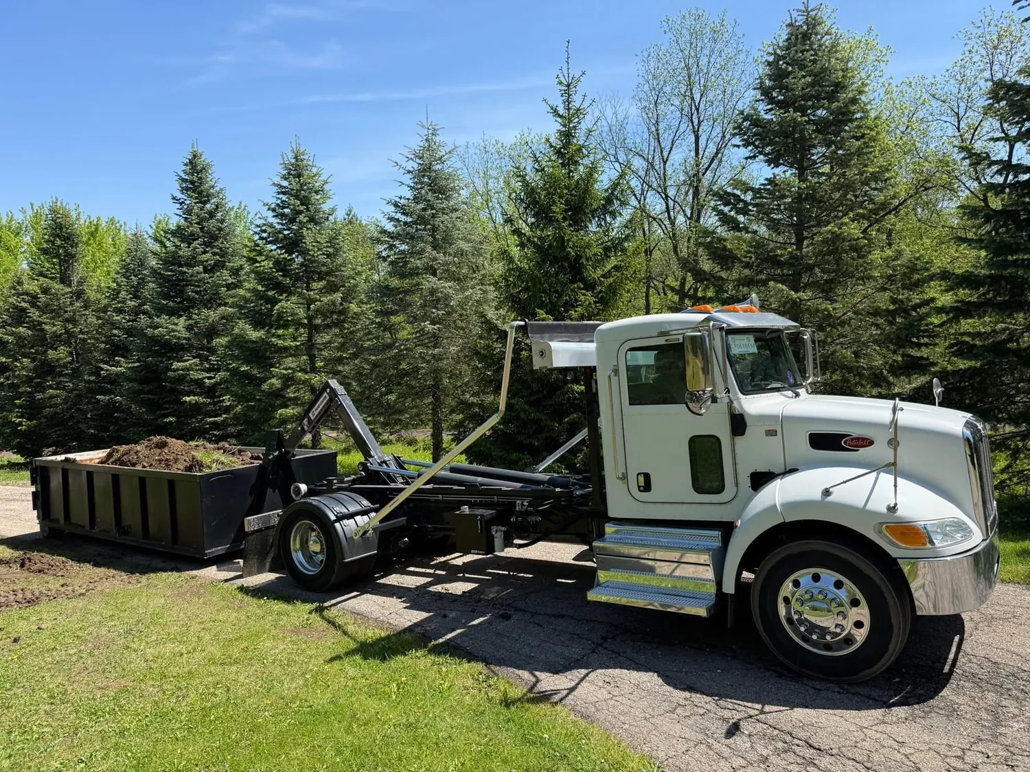 Roll-off truck delivering an empty dumpster onto a residential driveway between evergreen trees