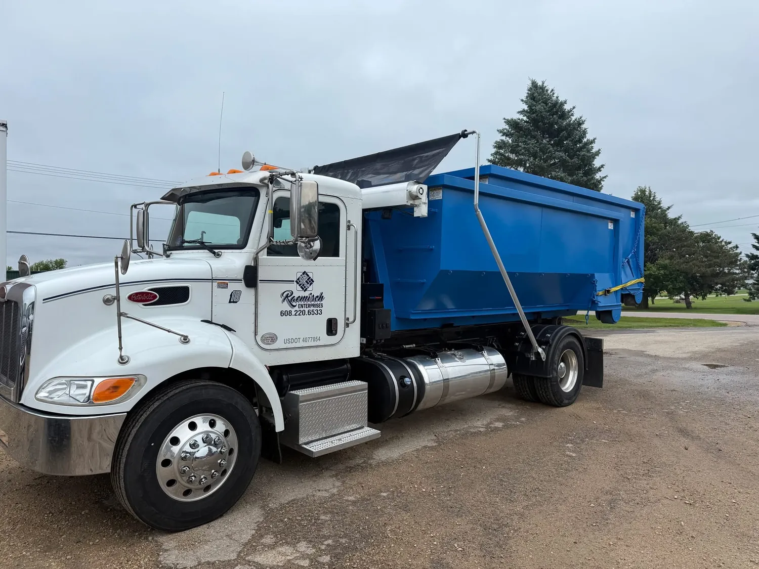 Roll-off truck with a loaded blue dumpster parked in a gravel lot on an overcast day